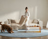 Woman in white loungewear styling a cream modular sofa with soft upholstered fabric, cosy knitted throw, dog, and glass coffee table in a modern living room.