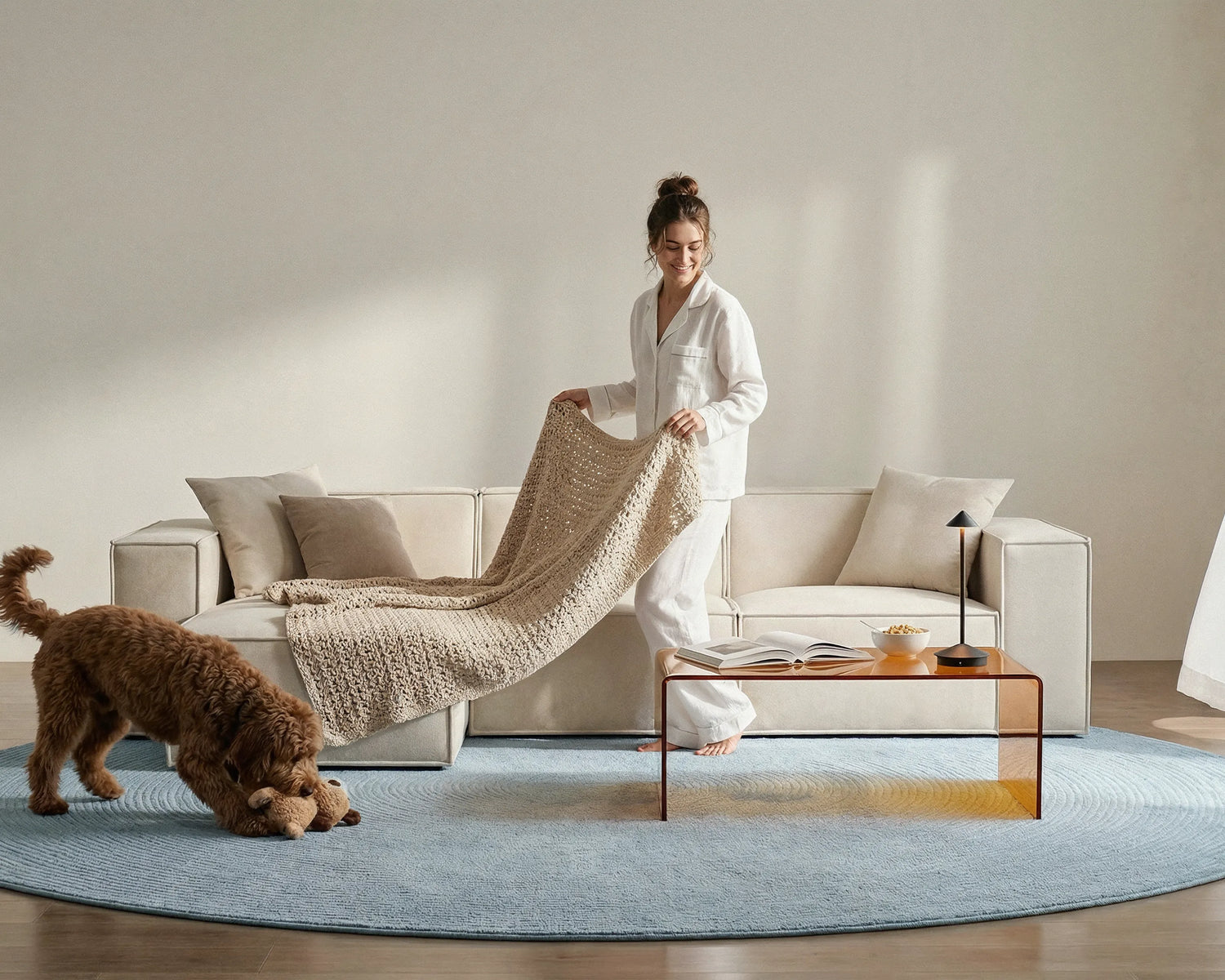 Woman in white loungewear styling a cream modular sofa with soft upholstered fabric, cosy knitted throw, dog, and glass coffee table in a modern living room.